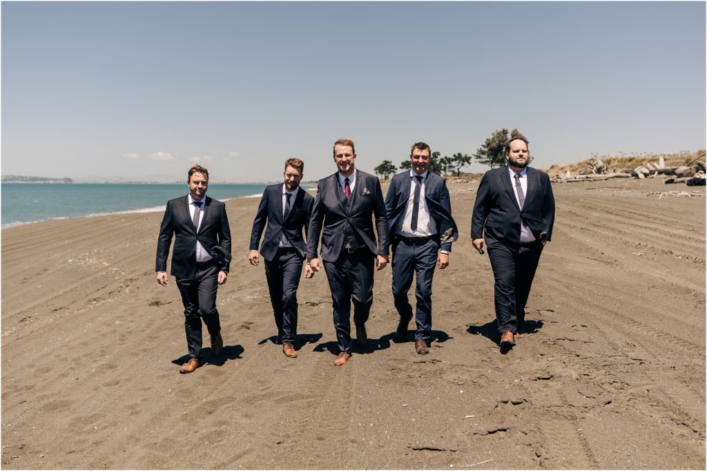 groom groomsmen walking on beach in navy suits in napier hawkes bay new zealand wedding summer sunny day