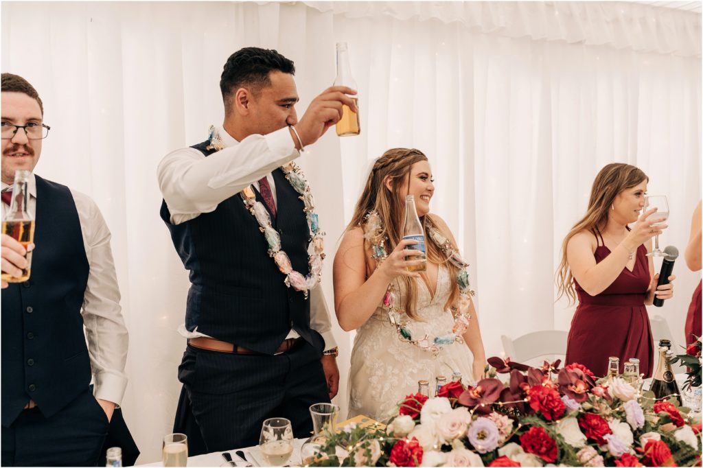 bride groom cheers at wedding reception holding long white and beer maroon flowers candy wreath around neck hideaway marquee wedding invercargill winton nz