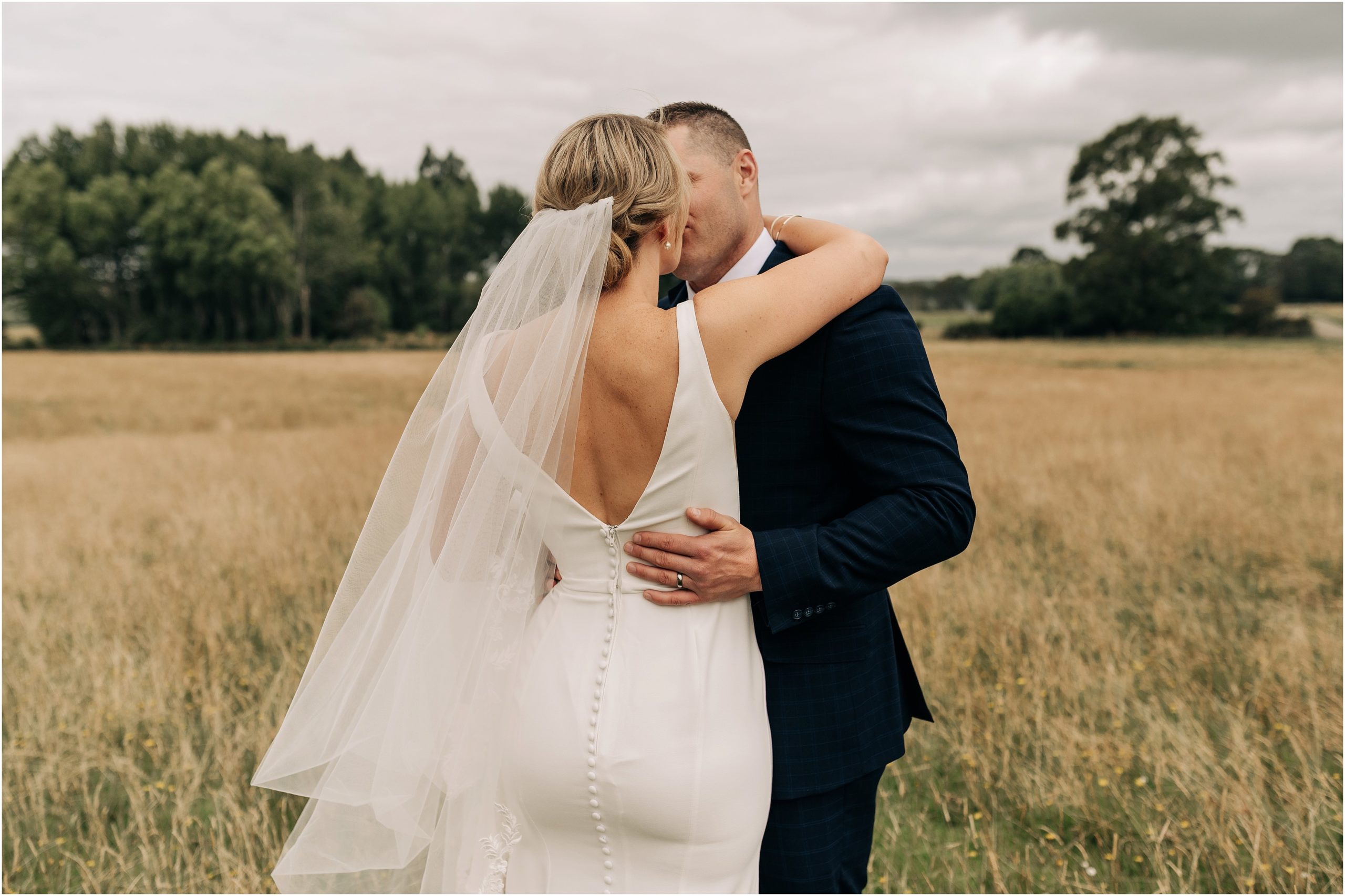 bride and groom in field of long grass christchurch new zealand wedding veil and black suit