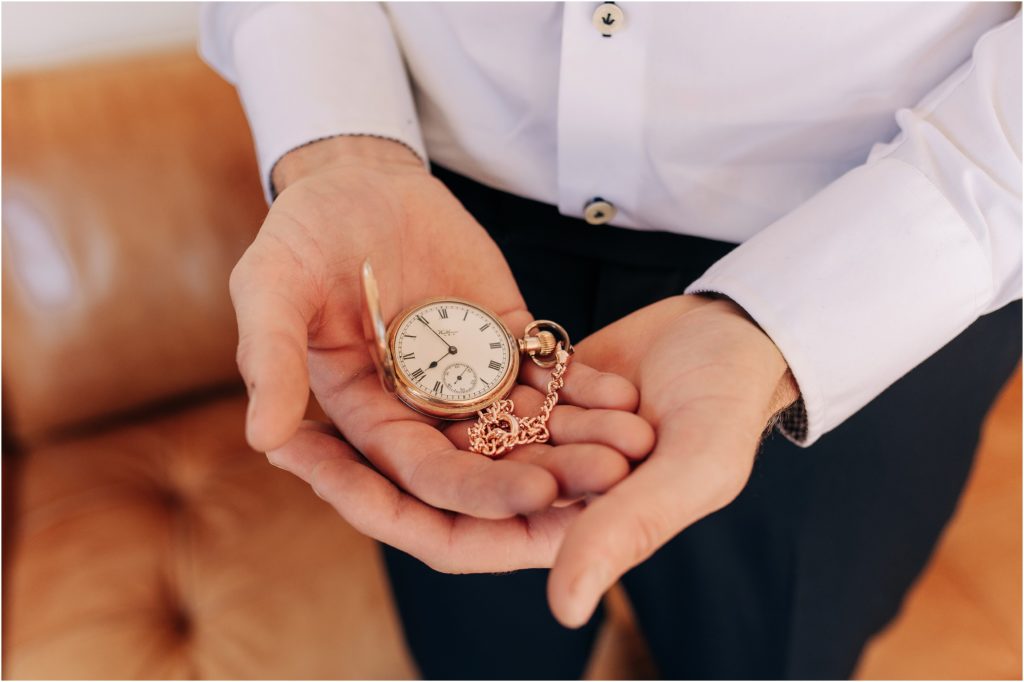 groom holding old vintage pocket watch gold on wedding day with white shirt navy pants brown leather couch new zealand christchurch