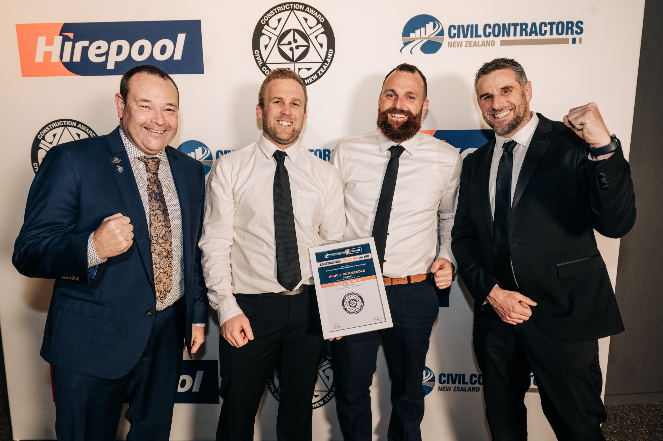 four men celebrate winning an award as they wear suits in front of a logo wall at a christchurch conference